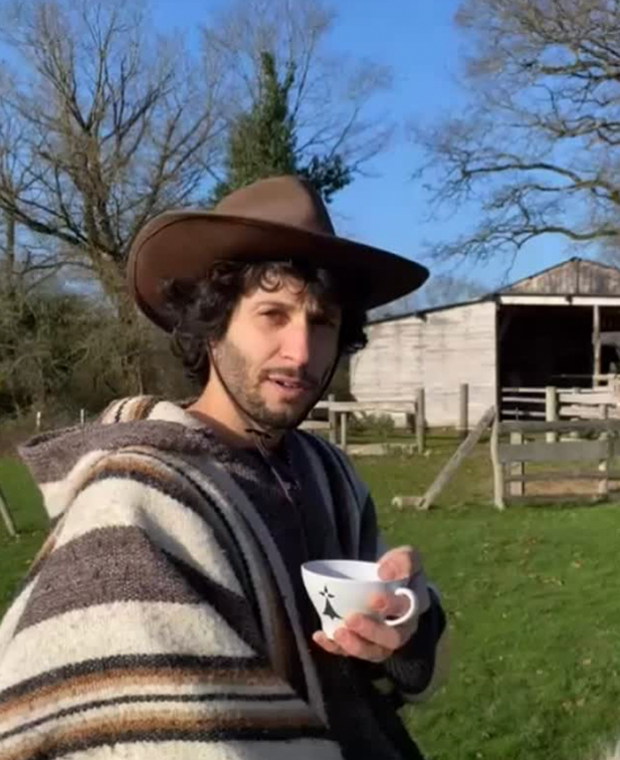 Un homme en poncho et chapeau, une tasse à la main, devant une grange